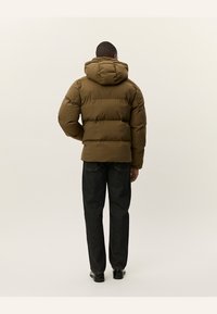 Chaqueta acolchada marrón con capucha, relleno grueso y corte rectangular. Combinada con jeans de mezclilla oscuros y zapatos negros. Vista trasera.