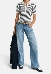 Polo w paski z kołnierzykiem, jasnoniebieskie spodnie typu wide-leg, wąski pleciony pasek w kolorze brązowym i ciemne buty, z ciemną kurtką zarzuconą na jednym ramieniu.