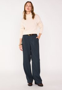 Pull à col roulé beige, coupe ample ; pantalon ajusté bleu foncé à rayures fines ; ceinture marron ; le mannequin pose avec une main dans la poche, accessoires minimalistes.