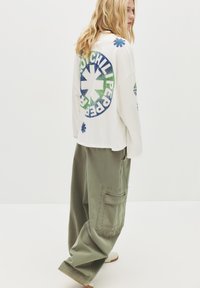 Blond persoon die zijwaarts kijkt, gekleed in een witte lange mouwen shirt met een groen en blauw Red Hot Chili Peppers logo en loszittende olijfgroene broek.