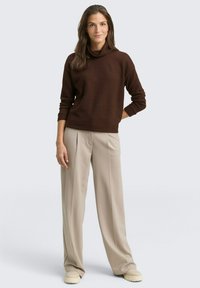 Un pull à col cowl marron en tissu doux, associé à un pantalon large beige clair. La tenue présente des coupes décontractées et un minimum de détails.