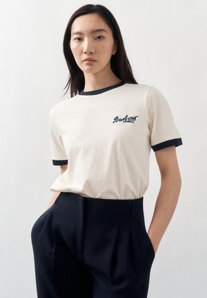 Młoda kobieta nosząca kremowy t-shirt Barbour z czarnymi lamówkami i granatowe spodnie z wysokim stanem, stojąca z rękami w kieszeniach na jednolitym tle.
