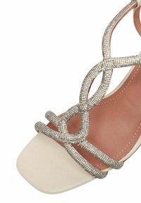Sandalo beige con cinturini, caratterizzato da scintillanti accenti argentati e una punta quadrata. Texture liscia con cinturini intrecciati decorativi.