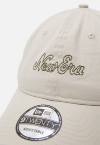 Bežs cepure ar regulējamu siksniņu, ar izcilu zaļo "New Era" logo. Ir apļveida uzlīme, kas norāda uz "9TWENTY" stilu. Klasisks dizains.