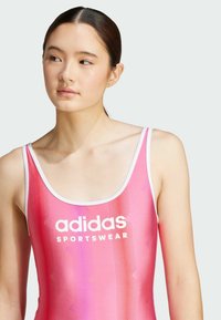 Růžový sportovní plavky s bílými ramínky a logem; obsahují texturovaný design s jemnými vzory a hladkým povrchem.