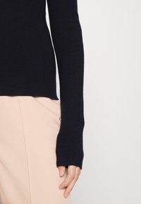 Maglione a coste navy con maniche lunghe, dotato di fori per il pollice e un orlo aderente, abbinato a pantaloni beige chiaro.