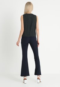 Svart, ärmlös topp med ett texturerat mönster och detaljer med nyckelhål i ryggen; matchad med marinblå utsvängda byxor; modellen bär vita blockklackade sandaler.