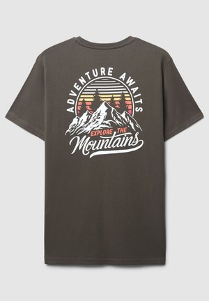T-shirt grigio scuro con un design grafico di montagne, alberi e un sole che sorge, accompagnato dal testo "L'avventura ti aspetta, esplora le montagne".