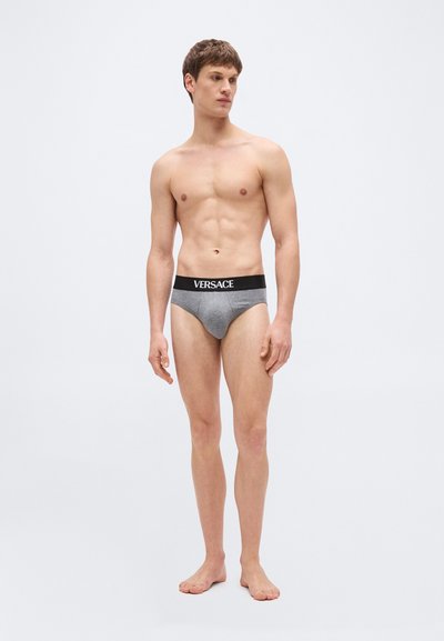 Γυμνός άνδρας που φοράει γκρι εσώρουχα Versace με μαύρη μέση, στέκεται μπροστά σε απλό ανοιχτό γκρι φόντο.