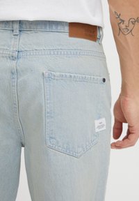 Jeans de denim azul claro con una etiqueta de marca de cuero, bolsillo trasero estándar y una pequeña etiqueta. La textura es suave y ligeramente desgastada.