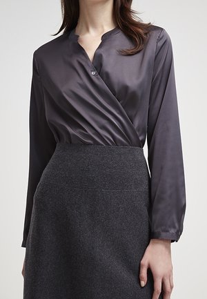 Femme portant un chemisier portefeuille en satin gris foncé rentré dans une jupe en laine gris foncé taille haute, avec des manches longues et un seul bouton visible.