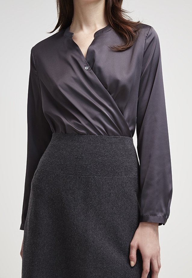 Femme portant un chemisier portefeuille en satin gris foncé rentré dans une jupe en laine gris foncé taille haute, avec des manches longues et un seul bouton visible.