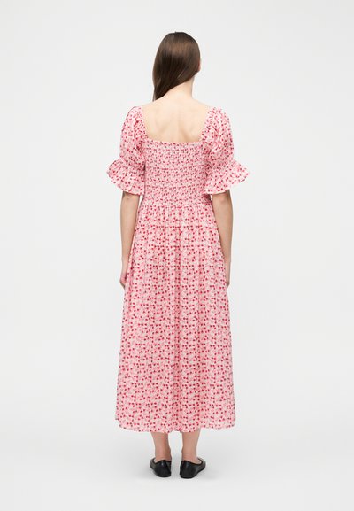 Γυναίκα που φορά ροζ μεσοφόρεμα με λουλούδια, φουσκωτά μανίκια και τετράγωνο ντεκολτέ, βλέποντας από πίσω, συνδυασμένο με μαύρα επίπεδα παπούτσια.