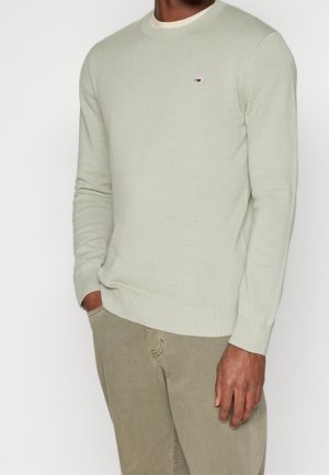 Homme portant un pull léger vert clair à col rond avec un petit logo sur la poitrine et un pantalon beige, debout avec une main dans la poche.