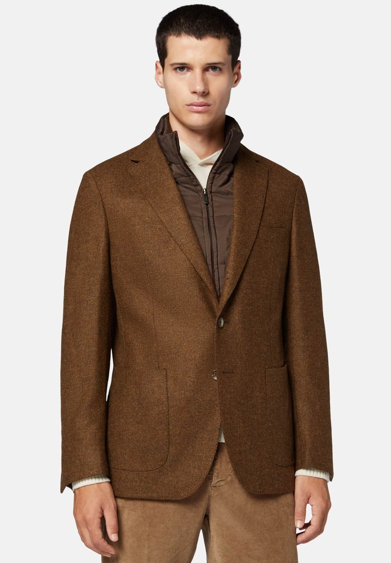 Blazer in tweed marrone con rever a scollo a V, chiusura a bottone singolo e due tasche frontali, indossato sopra un gilet imbottito marrone. Stile casual.