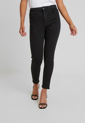 Nainen, joka kävelee mustuissa kapeissa farkuissa ja mustissa avokkaissa vaalean harmaan taustan edessä.