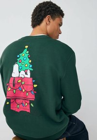 Zöld pulóver ünnepi Snoopy mintázattal, díszített karácsonyfával és színes fényekkel egy rózsaszín kutyaházon.