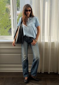 Femme portant un polo bleu clair, un jean délavé, des chaussures noires et des lunettes de soleil, portant un grand sac cabas noir texturé à l'intérieur près d'une fenêtre.