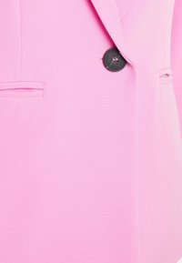 Felroze blazer met een enkele zwarte knop die bij de taille is vastgemaakt en twee horizontale schuine zakken aan de voorkant.