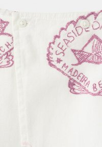 Tissu blanc avec texte brodé en violet indiquant "SEASIDE" et "MADEIRA BEACH" autour d'un motif de coquillage près de deux boutons blancs.