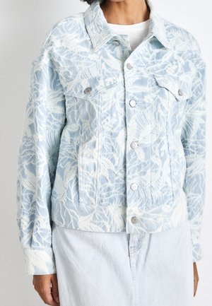 Femme portant une veste en jean bleu clair avec broderie florale blanche, boutons métalliques et poches poitrine à l'avant, sur une chemise blanche.