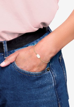 Guldtonet armbånd med en glat hvid stendetalje. Båret på et håndled, med et enkelt, delikat design mod denim og en lyserød top.