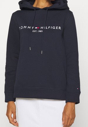 Granatowa bluza z kapturem z logo "Tommy Hilfiger EST. 1985" na wysokości klatki piersiowej, sznurkami oraz małą flagą na mankiecie lewej rękawa.