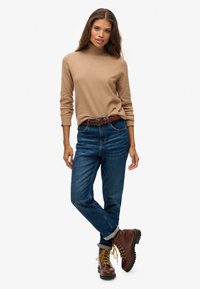 Donna che indossa un dolcevita beige, jeans blu con una cintura marrone e stivali con lacci marroni, in piedi con una gamba incrociata sopra l'altra.
