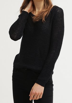 Schwarzer gestrickter Pullover mit langen Ärmeln, lockerer Passform und strukturiertem Muster. Kombiniert mit schwarzen Hosen, die einen lässigen Look zeigen.