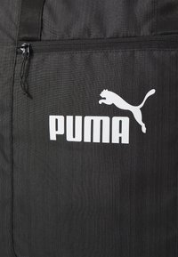 Zwarte stoffen tas met een wit Puma-logo. Het heeft een ritsvak en een gestructureerd oppervlak, ontworpen voor duurzaamheid en functionaliteit.