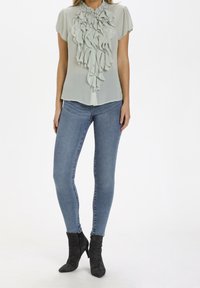 Blouse vert clair avec un devant volé, manches courtes, portée avec un jean skinny bleu et des bottines noires. Tissu lisse et coupe ajustée.