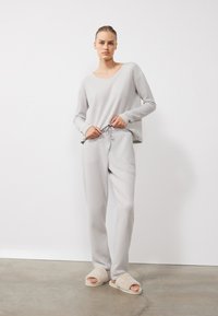 Sivi komplet loungewear od rebraste tkanine koji se sastoji od majice s V-izrezom i dugih rukava te hlača svezanih u struku, upotpunjen bež plišanim papučama.