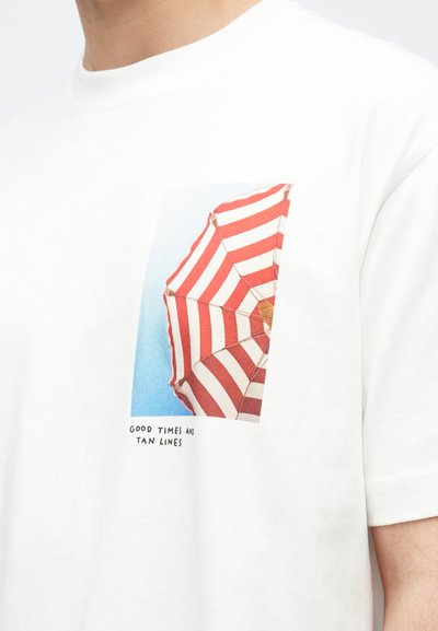 Λευκό βαμβακερό μπλουζάκι με τυπωμένο γραφικό που απεικονίζει μία κόκκινη και λευκή ριγέ ομπρέλα παραλίας πάνω σε μπλε φόντο με κείμενο.