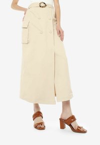 Beige wikkelrok met dubbelzijdige knopen en zijzakken, gecombineerd met bruine sandalen met hakken met geweven bandjes.