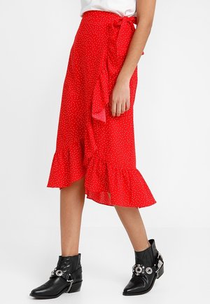 Femme portant une jupe portefeuille rouge à pois avec des volants et un nœud noué, associée à des bottines noires cloutées, debout devant un fond blanc.