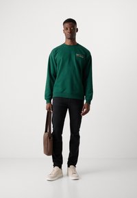 Sweatshirt vert avec col rond, détails brodés ; associé à un jean noir et des baskets beiges. Sac marron avec bandoulière.