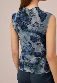 Ærmeløs top i et blåt tie-dye mønster med blomsterdesigns. Blødt stof med en subtil glans og rå kanter ved halsen.
