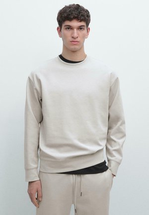 Junger Mann, der nach vorne blickt, trägt einen beigen Pullover und passende Jogginghosen mit einem schwarzen Hemd darunter.