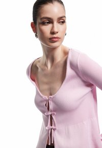 Cardigan a coste rosa con scollatura a V profonda, caratterizzato da tre chiusure con fiocchi sul davanti e una sezione inferiore a texture contrastante.