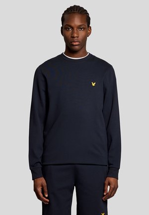 Giovane uomo con i capelli intrecciati che indossa una maglia nera a maniche lunghe e pantaloni, con un piccolo logo giallo a forma di uccello su entrambi gli indumenti.
