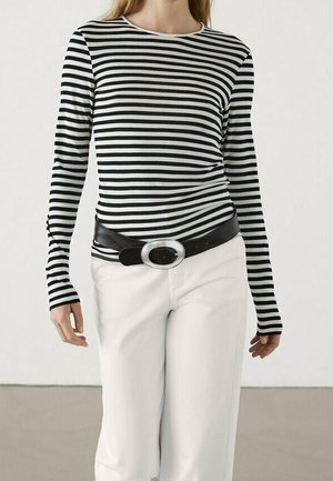 Femme portant une chemise à manches longues à rayures horizontales noires et blanches, un pantalon blanc et une ceinture noire avec une grande boucle argentée.