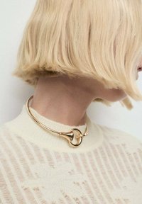 Collar de choker de metal dorado con un diseño curvo y elegante, y un acento central. Llevado sobre un suéter crema claro y texturizado.