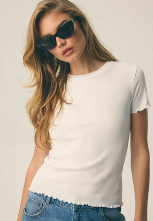 Kobieta o długich blond włosach, nosząca czarne okulary przeciwsłoneczne w kształcie kociego oka, biały prążkowany t-shirt z falbankami na krawędziach i niebieskie dżinsy z denimu.