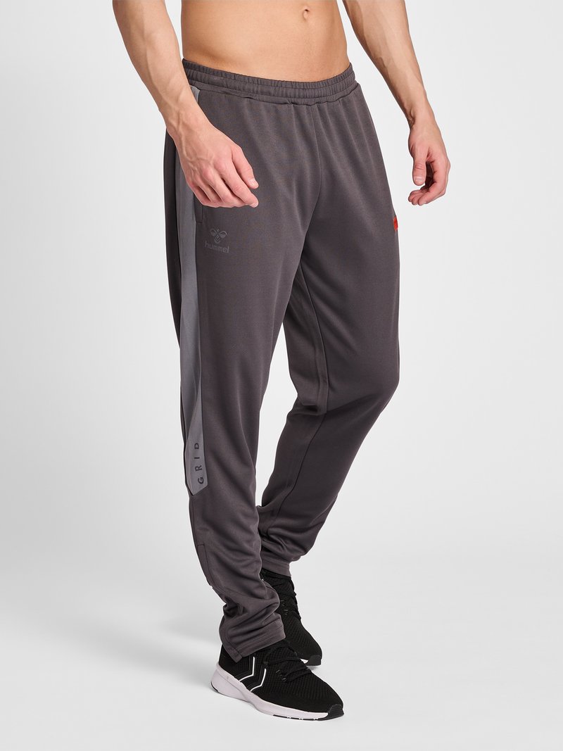 Torso et jambes d’homme portant un pantalon de sport gris foncé avec une ceinture élastique et des chaussures de sport noires, debout sur un fond blanc.