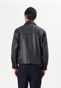 Jachetă din piele neagră cu guler ascuțit, detalii cu nasturi pe manșete și croială dreaptă. Prezintă o textură netedă și un design minimalist.