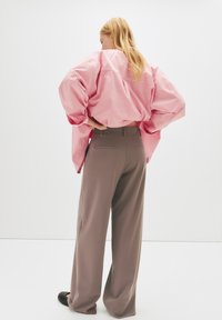 Lyserød stribet oversized skjorte med brede ærmer, parret med løse brune bukser. Enkel, minimalistisk design, sat mod en ensfarvet baggrund.