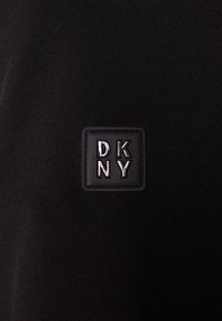 Svart tyg med en slät textur som har en fyrkantig DKNY-logotyp i svart med upphöjd text. Logotypen är centralt placerad.