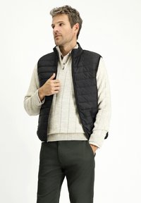 Gilet nero trapuntato con colletto alto, indossato sopra un maglione grigio chiaro a coste. Il modello è in piedi, con una mano sul gilet, indossando pantaloni verde scuro.