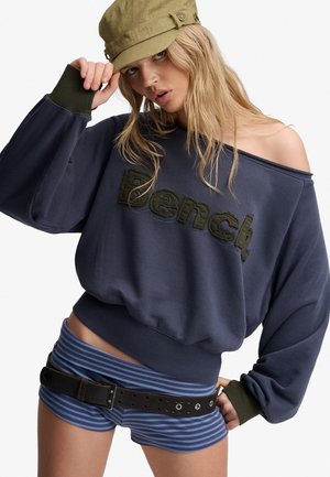 Jeune femme portant un sweat-shirt ample bleu marine avec le logo "Bench", un short bleu rayé avec une ceinture noire et une casquette kaki, posant avec une main sur la casquette.