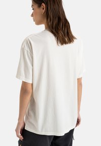 Jasno beżowa koszulka z krótkim rękawem o luźnym kroju, miękka w dotyku, wykonana z bawełny, z okrągłym dekoltem. Tylny widok pokazuje gładki materiał bez detali.